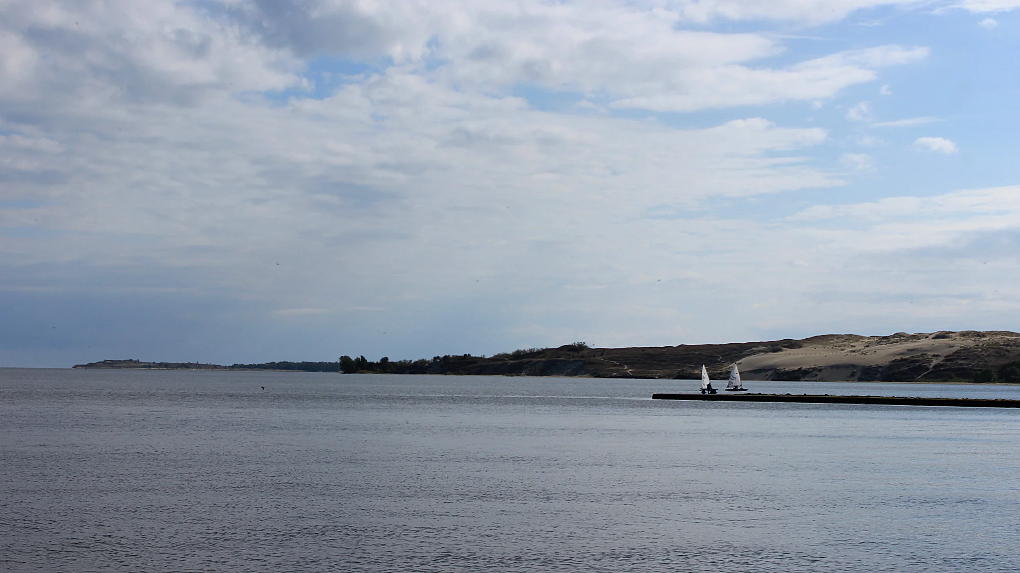 The empty Baltic Sea from Nida with a dock, two sailing ships and a dune in the distance.