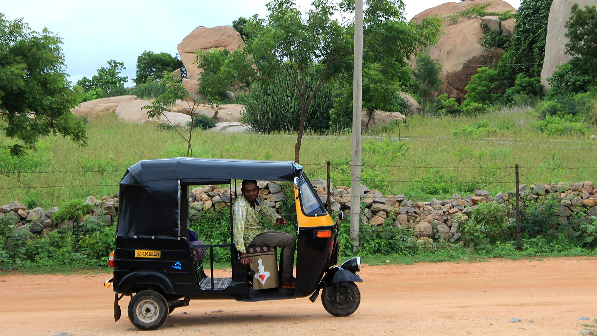 A black and yellow rickshaw parked on a hill with the driver looking at the camera. Photographed from the side.