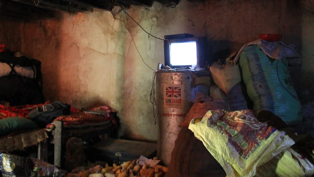 A dark Nepalese village room with a small television and corncobs on the floor.