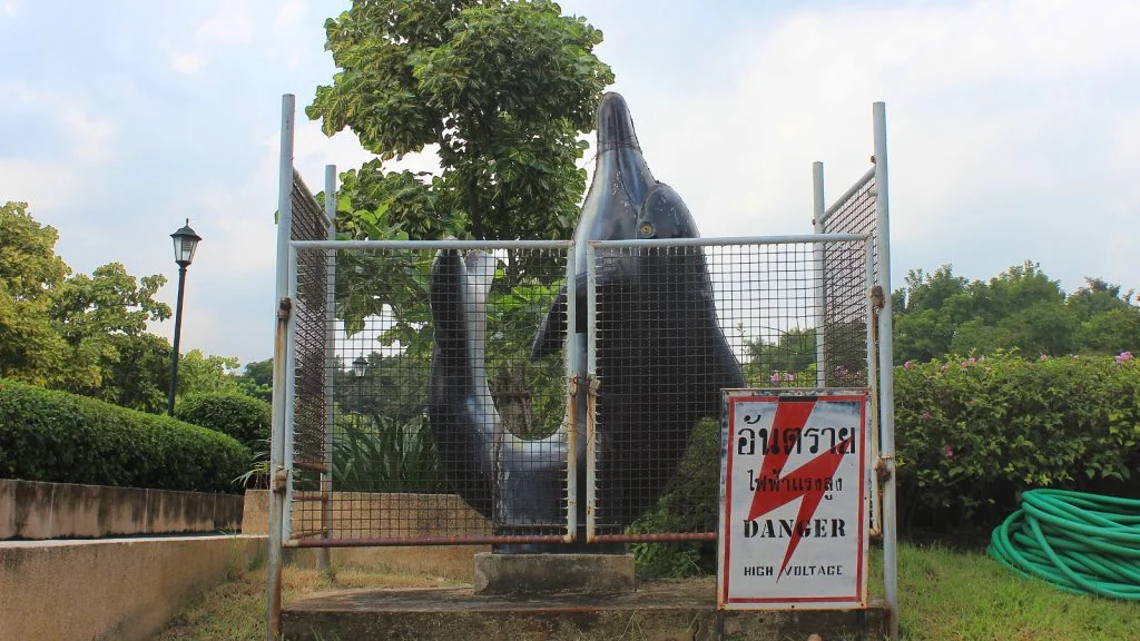 A fenced dolphin statue with a "DANGER HIGH VOLTAGE" sign on the metal fence in a park in Silom, Bangkok.