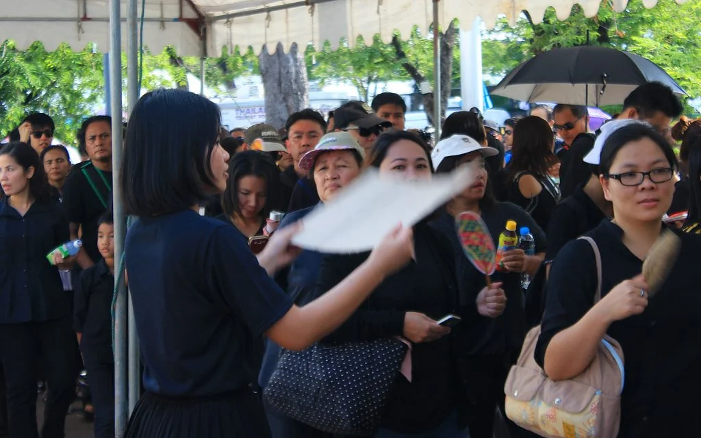 A Thai woman waving a cardboard fan to cool people waiting on a line to enter the Grand Palace in Bangkok, Thailand. Mourners wait to pay their respects for late King Bhumibol.
