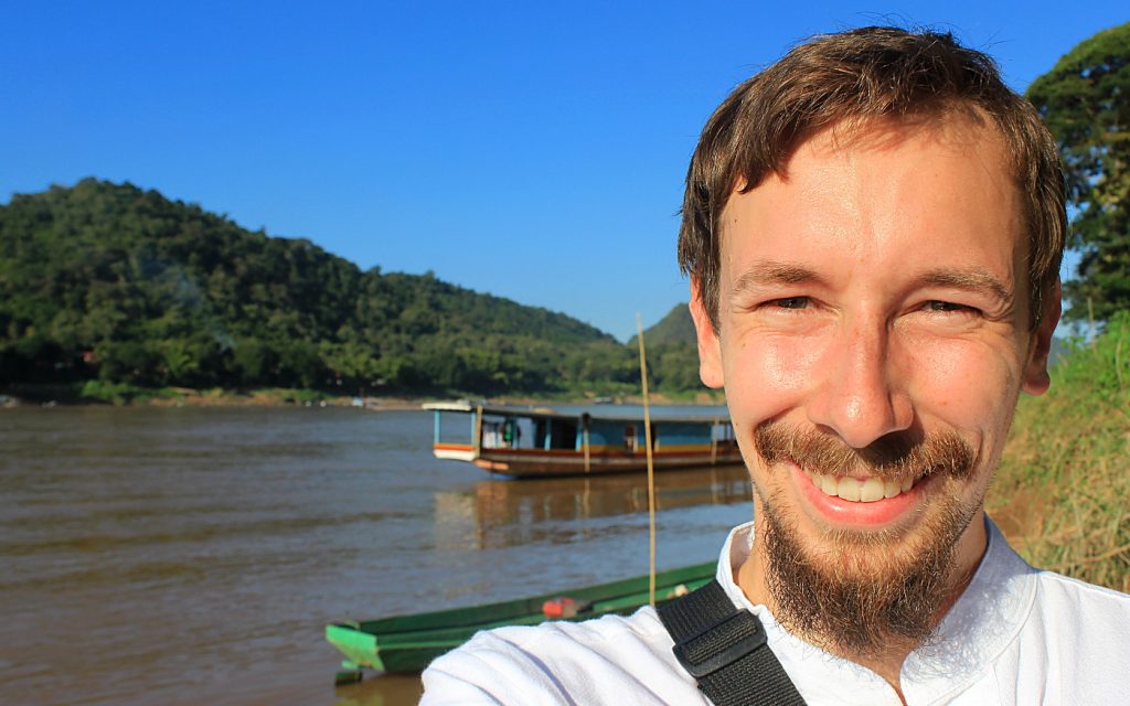 Arimo Koo travels. RTW travel blogger taking a selfie by Luang Prabang dock with a slowboat in the background.